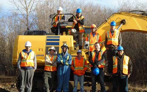 A crew standing in front of an excavator and other equipment at a bush worksite surrounded by spruce and poplar.
