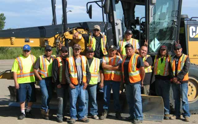A crew of thirteen trainees in high-visibility vests standing together in front of a Caterpillar wheel loader on a gravel yard.