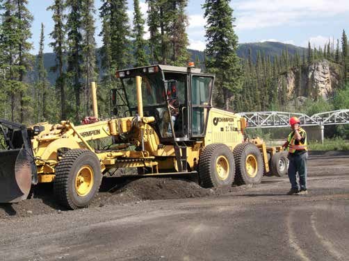 A motor grader working a newly built gravel road cut through forest, with a second machine behind it.