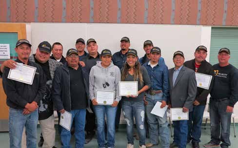 Fifteen graduates standing together indoors, several holding certificates, at the end of a heavy equipment operator program.