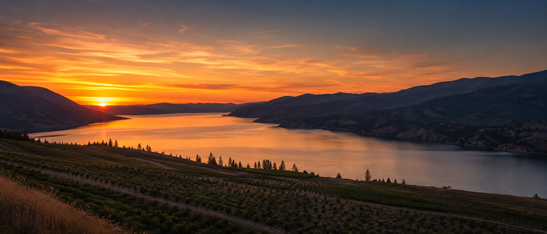 Sunset over a long lake in the Okanagan valley, with orchard benchland on the near slope and silhouetted hills beyond.